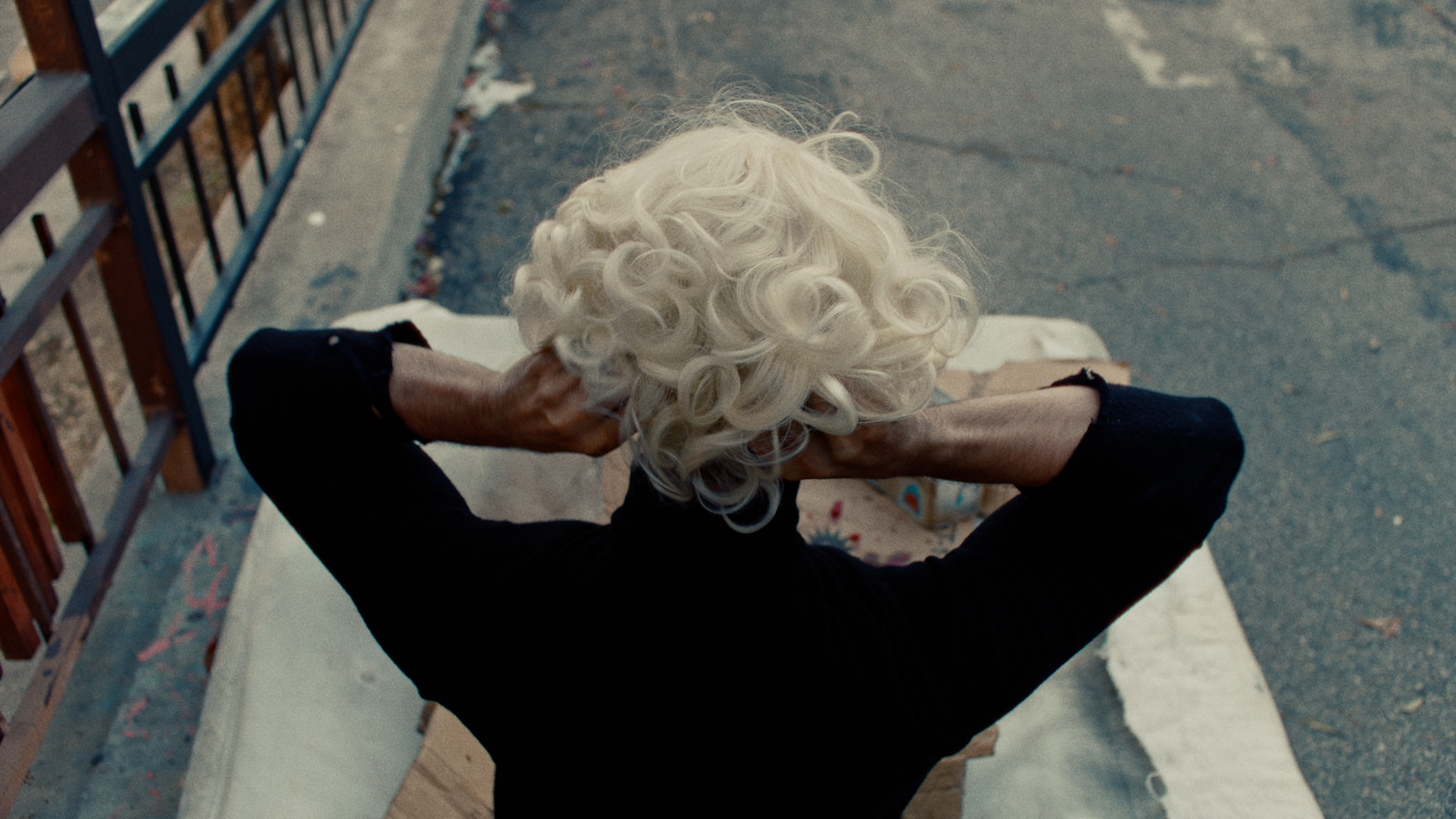A figure adjusts a platinum blonde wig, shot from above on a gritty curb